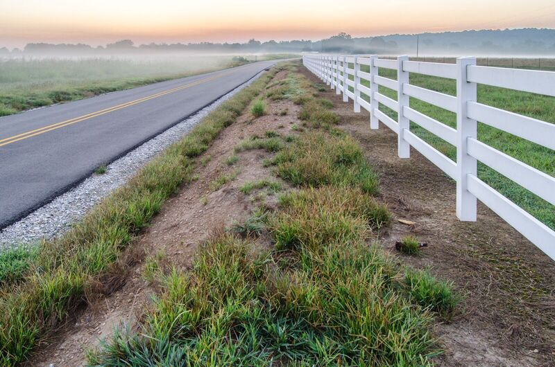 White vinyl four-rail ranch fence along a country road in morning fog
